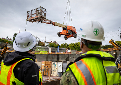Two shipyard workers standing in front of a crane being suspended in the air.